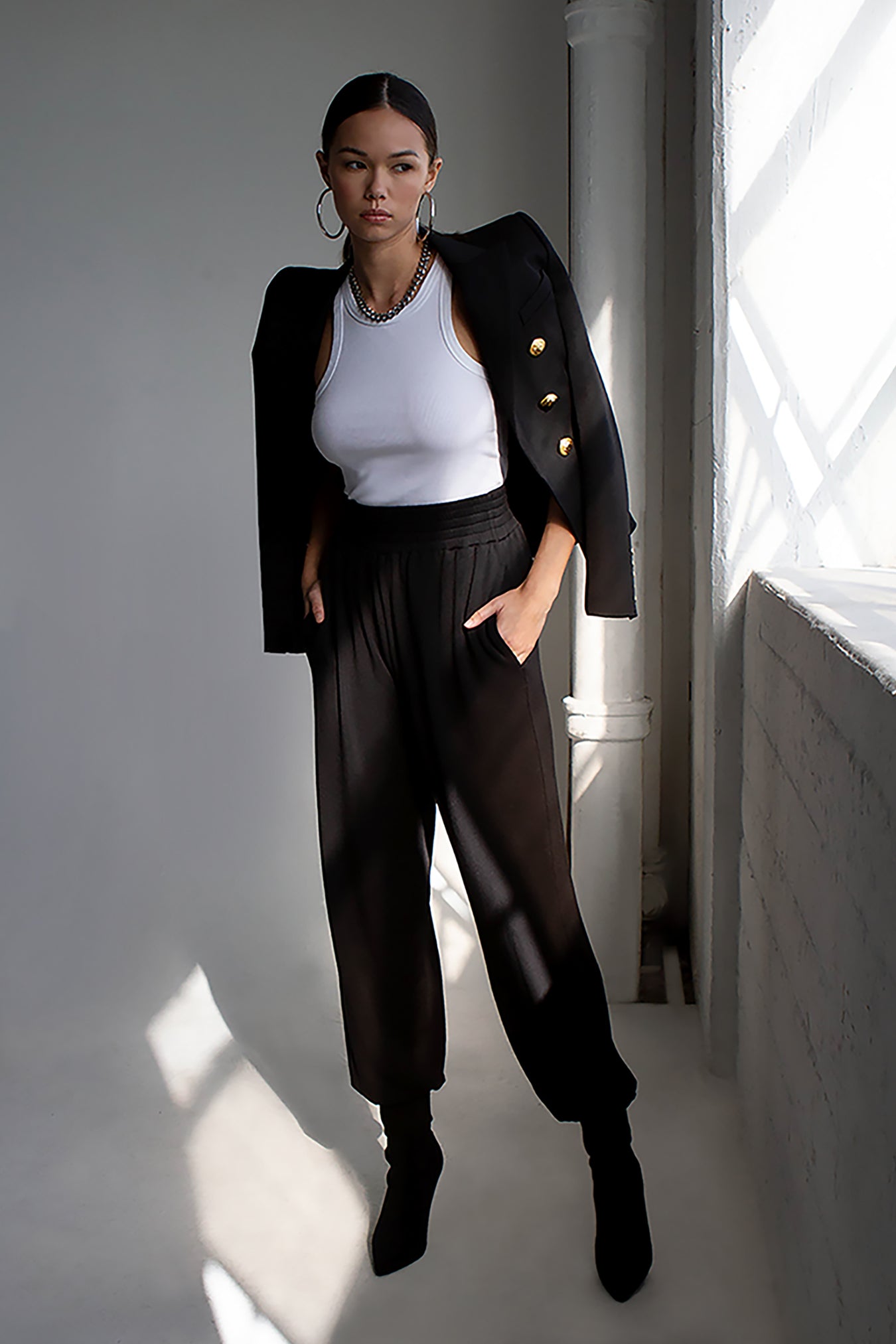 Hands in her pockets, a model standing next to a window, wearing a black blazer, a white racer tank top, and a pair of high waisted black fleece sweatpants with black sock booties. 