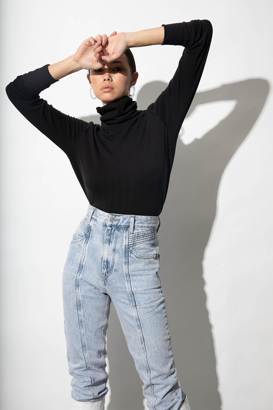 A model with her arms up and hands resting on her forehead is wearing a black turtleneck and blue acid-wash jeans.