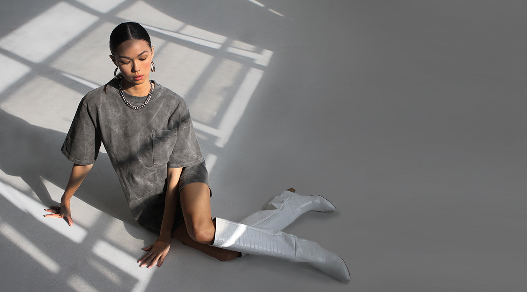 A model sitting on the floor in a partially sunlit room is wearing a vintage washed t-shirt dress with silver hoop earrings, a silver chain link necklace, and a pair of white knee-high boots